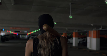 Naughty girl in a balaclava holds a baseball bat in her hands and poses on the territory of a car parking. Cheeky sexy woman in mask shows disrespect and aggressionの写真素材