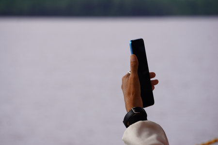 A man's hand holding a cellphone with a blurry sea background