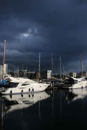 Waterfront marina and surrounding buildings in Ipswich, Suffolk, United Kingdom with very dark storm clouds and sunlight illuminating the boats and buildings.の写真素材