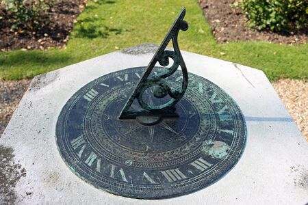 Metal sundial on stone plinth with gnomon sharply in focus with background of flower beds and lawn...