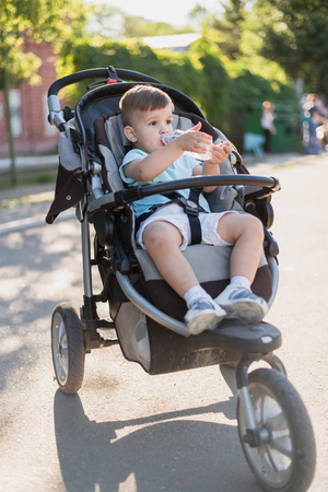 cute baby sits in a stroller and drinks clean water and on a hot summer dayの写真素材