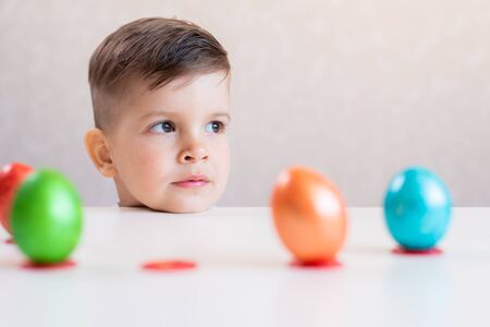 A little beautiful cute boy laid his head on a white table on which in a row stand multi-colored...