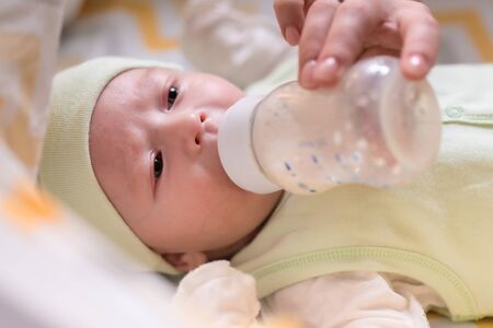 young mother feeds her newborn baby milk from a bottle that lies in the bed
