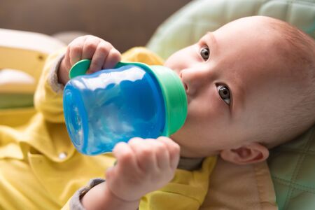 The infant drinks water from a bottle lying in a high chair for feeding.