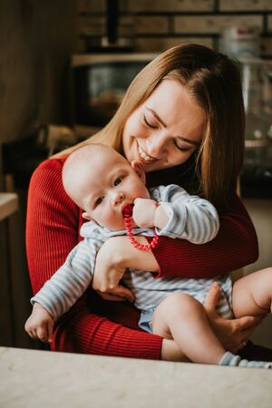 A mom holds a baby in her arms. Mother hugs her toddler. A young woman has fun and rejoices with the child. A girl clutches a infant.の写真素材