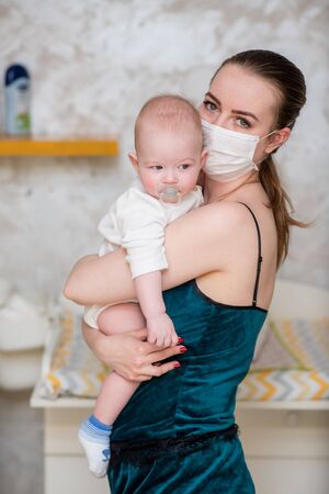 A mother in a protective mask with a baby is sitting at home in quarantine during the coronovirus and covid-19 pandemic.の写真素材