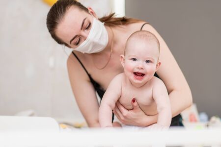 A mother in a protective mask with a baby is sitting at home in quarantine during the coronovirus and covid-19 pandemic.の写真素材