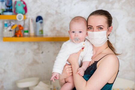 A mother in a protective mask with a baby is sitting at home in quarantine during the coronovirus and covid-19 pandemic.の写真素材