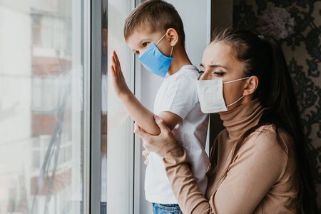 A woman with a child in medical masks is sitting at home in quarantine and looking out the window. Prevention of Coronovirus and Covid -19の写真素材