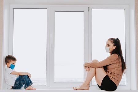 mother with a child in medical masks are sitting on the window at home in quarantine and looking out the window. Prevention of Coronovirus and Covid -19の写真素材