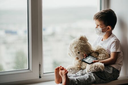 A small child sits at home with a teddy bear in quarantine and plays on the phone. Prevention of coronavirus and Covid - 19の写真素材