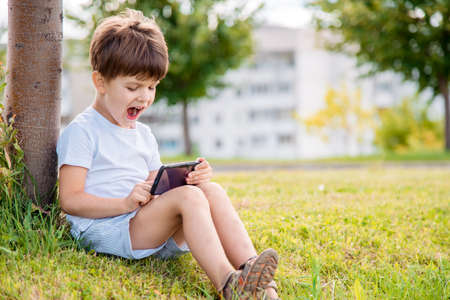 Cheerful boy sitting on the grass looks cartoons in the phone in the summer at sunset. Cute baby...