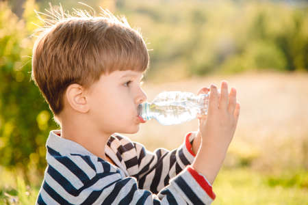 Cute boy sitting on the grass drinks water from a bottle in the summer at sunset. Child quenches thirst on a hot dayの写真素材