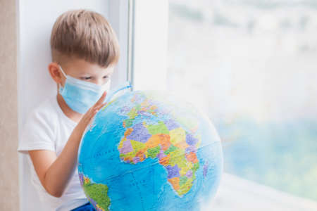 The masked kid sits at home in quarantine. The child is examining the globe while sitting on the...