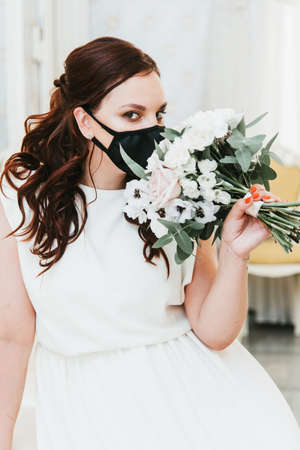 Portrait of a beautiful bride with a bouquet in a medical protective mask on her face.Wedding during the coronavirus period.の写真素材