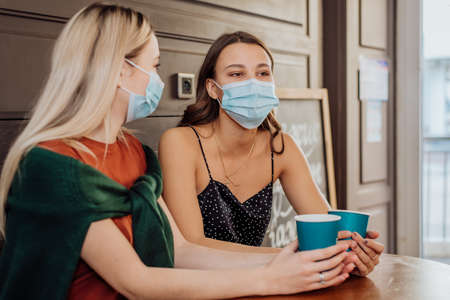 Two beautiful girls with masks on their faces spend time together in coffee and drinking tea. Protection against coronavirus Covid-19 during the pandemicの写真素材