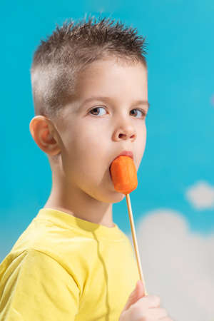 A small child eats a carrot on a stick. Boy gnaws vegetables on a blue background.