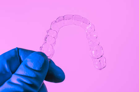 Invisible dental braces are held by a hand in a blue glove on a pink background. Plastic braces dentistry retainers to straighten teeth.の写真素材