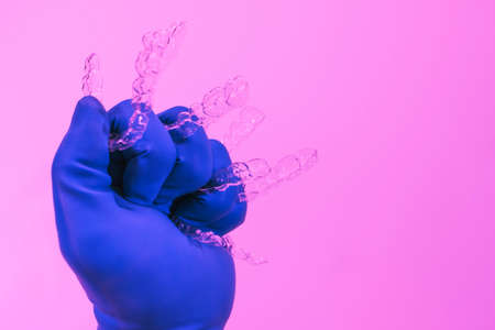 A hand in a blue glove holds aligners for aligning teeth on a pink background. Plastic braces dentistry retainers to straighten teeth.の写真素材