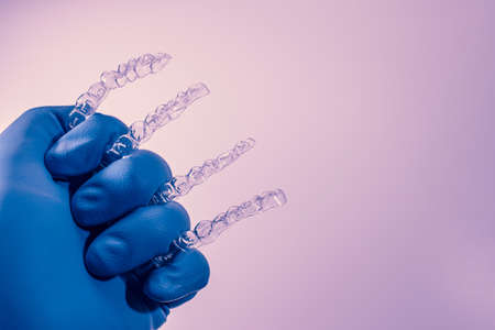 A hand in a blue glove holds aligners for aligning teeth on a purple background. Plastic braces dentistry retainers to straighten teeth.の写真素材