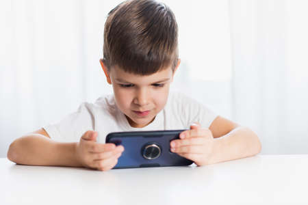 A cheerful child in a white T-shirt plays games on the phone at home. Happy boy looks at his smartphone.の写真素材