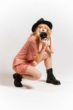 Portrait of a beautiful woman photographer in a hat posing with a camera in her hands in the studio on a white backgroundの写真素材