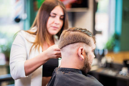 Hairdresser woman cutting hair with clipper bearded man in barbershopの写真素材