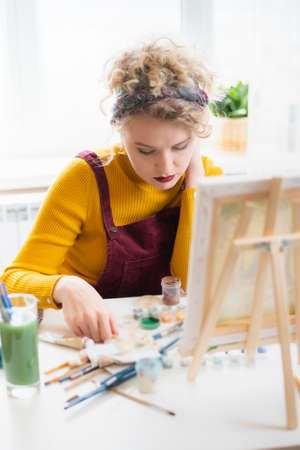 Beautiful woman artist sits in front of a painting and paints with brushes on canvas at home