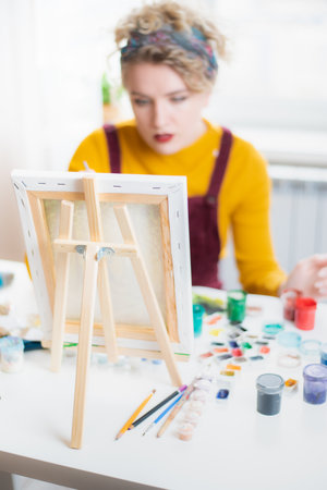 Beautiful woman artist sits in front of a painting and paints with brushes on canvas at home