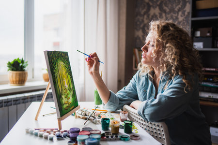 Beautiful woman artist sits in front of a painting and paints with brushes on canvas at homeの写真素材