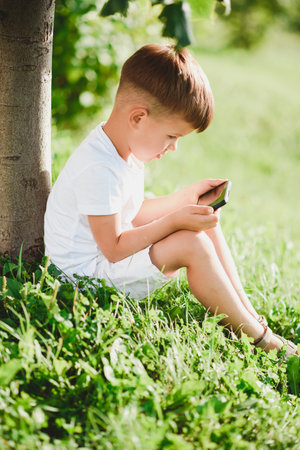Cheerful boy sitting on the grass looks cartoons in the phone in the summer at sunset. cute baby having fun in natureの写真素材