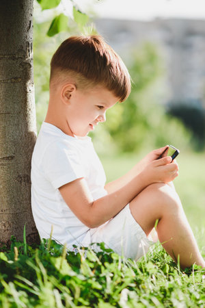 Cheerful boy sitting on the grass looks cartoons in the phone in the summer at sunset. cute baby having fun in natureの写真素材
