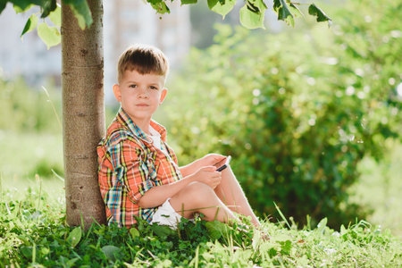 Cheerful boy sitting on the grass looks cartoons in the phone in the summer at sunset. cute baby having fun in natureの写真素材