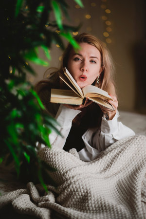 Young woman reading a book while sitting on her bed at home at nightの写真素材