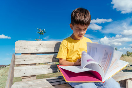 Boy reading a book sitting on a bench in nature during the dayの写真素材
