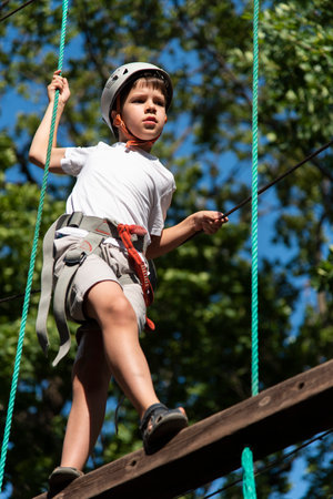 A boy in a rope park in nature learns to rock climb. A child in a helmet walks along a ropeway...