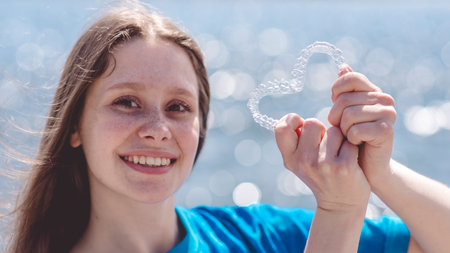 Dental care. Smiling girl holding heart-shaped aligners. Plastic retainers for teeth alignment.