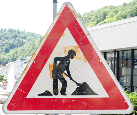 A rusty triangular sign warning of roadworks ahead with a modern city building in the backgroundの写真素材