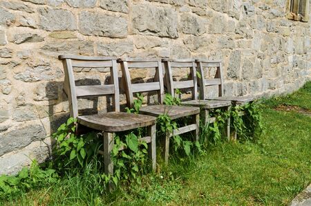 4 old wooden chairs in overgrowing grass and ivy in front of a stone church wal.lの写真素材