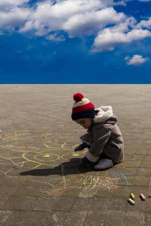 Single girl aged 4 in hat drawing with chalk under bright blue skyの写真素材