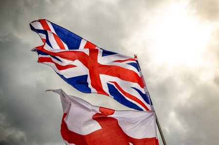 English Union Jack and saint George flag flying in the wind against a grey cloudy sky