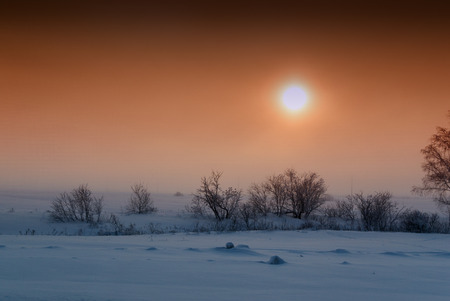 Setting sun with an orange cloudy sky settles over fresh fields of snow interspersed with bushes and trees in this winter style landscape image.の写真素材