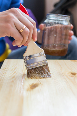 Casual clothed young woman holds a jay of home DIY varnish and uses a paintbrush to paint a pine chair or table surface. Lifestyle design activity with copy space area for home improvement use.の写真素材