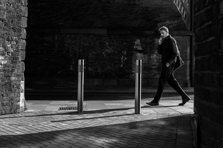 CHELMSFORD - ENGLAND 20TH APRIL 2016 - Man walks though a sunlit area as he approaches a bridge. He is talking on his mobile phone in Chelmsford England in April 2016のeditorial素材