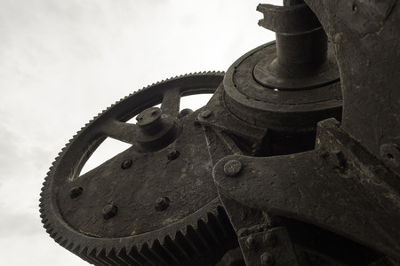 Single black painted cog wheel from an bygone industrial era set against a grey sky.