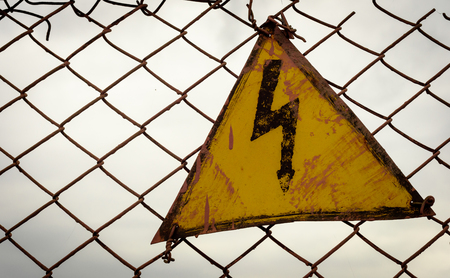 Lightning bolt electricity warning sign in a yellow triangle set against a dark sky wire mesh...