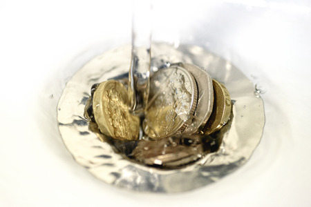 British money including a one pound coin stuck in a sink drain plughole as water flows over the coins.の写真素材