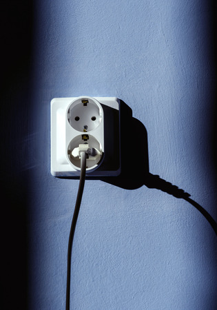 Electrical plug socket with electric cable inserted into one of the plugs with a blue wall background and sunlightの写真素材