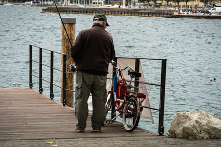 view of old man from behind with fishing rod and bicycle at lake Gardaのeditorial素材