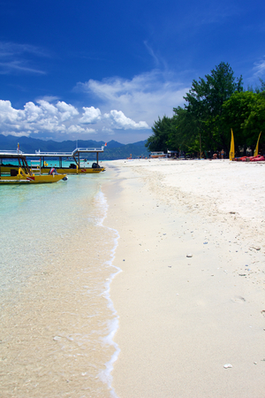 Beach on Gili Trawangan island, Indonesia, with yellow boats, white sand and small rock in foreground on sunny dayの写真素材